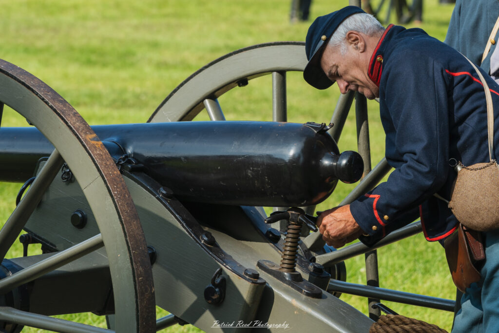A Union soldier preparing a cannon to fire, focusing intently on his task. Dressed in a blue uniform, he carefully loads the cannon, surrounded by fellow soldiers who assist with the operation. The scene captures the tension and urgency of wartime preparations on the battlefield.
